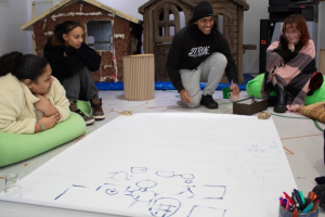 a group of people sitting on the floor around a large sheet of paper with drawings on it