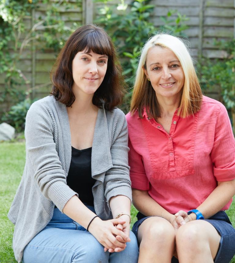 Two women sitting side by side, one with brown hair and wearing a grey cardigan and jeans, the other with blonde hair and a pink top and skirt 