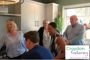 A mixed group of people standing in a kitchen, laughing and smiling. Croydon Fostering logo is in the bottom right corner of image.