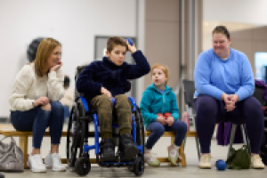 A young boy in a wheelchair about to throw a ball in a sports hall. Two adults and a child are sitting behind him watching and smiling.