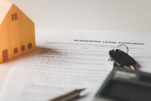 Stock image of a small wooden model of a house next to a rental lease agreement and a set of keys.