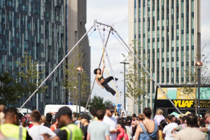 Aerial circus performer on rig performing above crowd in town centre with modern buildings in background.