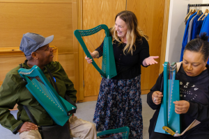 People taking part in a creative activity with small handheld harps in an indoor room, with one person demonstrating while two others play along nearby.