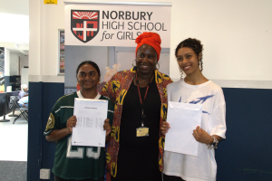 Two smiling students holding their exam results with a smiling teacher inbetween them