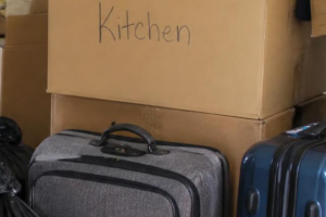 Suitcases and black bags next to stacked cardboard boxes with Kitchen written on one of them,